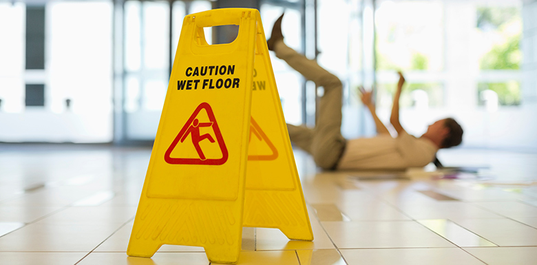 Inside of building entry, white tile floor with yellow caution wet floors sign. In the background a person fallen on the ground