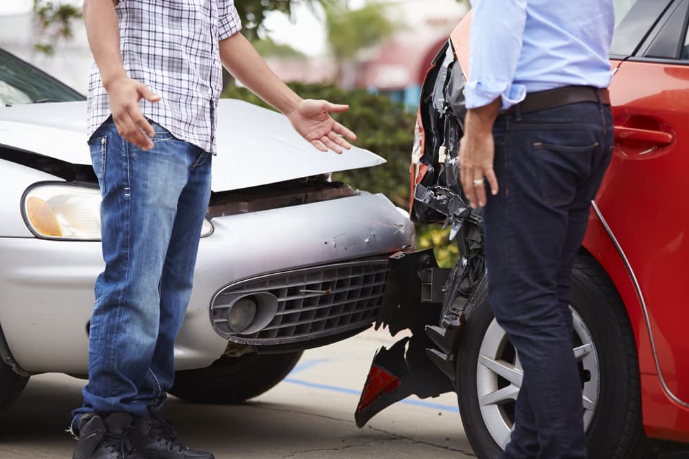 Two drivers arguing with each other after a traffic accident, standing next to their damaged vehicles on the roadside.